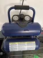 Front view of blue twin-tank Campbell Hausfeld compressor showing gauges and warning label