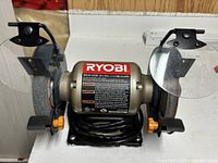 Front view showing RYOBI bench grinder with both grinding wheels, eye shields, and power cord