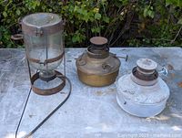Three items on table: brass heater, galvanized heater, glass bottle in iron frame