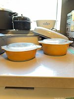 Front view showing two amber Pyrex bowls with lids, white enamel roaster, CorningWare dish, speckled pot and partial Le Creuset pot