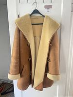 Full view of beige shearling coat hanging on hanger showing exterior suede and toggle closures