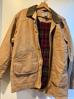 Full view of tan canvas jacket with corduroy collar and plaid lining displayed on hanger