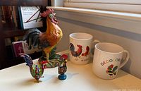 Group view of all rooster figurines and two mugs on tabletop