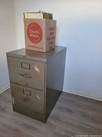 Front view of cabinet showing two drawers, handles, label frames and finish wear; box of folders on top