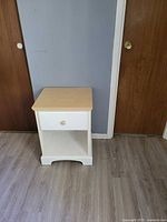 Front view of white nightstand showing wood top, one drawer with knob, and open lower shelf