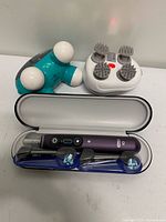 All three devices displayed together: toothbrush in open case, Homedics massager, white scalp massager