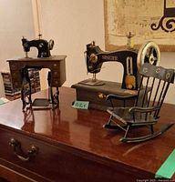 Front view of Singer toy sewing machine, unbranded treadle sewing machine and wooden rocking chair on tabletop