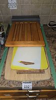 Stack of cutting boards on countertop showing wood slatted, plastic yellow, plastic white, large bamboo; metal mesh trays in background