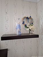 Shelf on wall holding angel figurine, silver cup with flowers; wicker wreath on wall