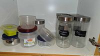 Interior of cabinet showing five tall clear glass jars with stainless lids and chalkboard labels beside stacks of plastic SnapTite and assorted food containers with coloured lids.