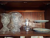 Shelf view showing pair of cut glass goblet vases, round cut glass platter, glass teapot warmer and two-tier dessert stand