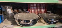 Two stainless steel pans with lids and a glass coffee carafe on shelf