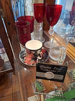 Group view of teacup, saucer, etched tankard, two cranberry goblets and cranberry tumbler displayed on glass shelf