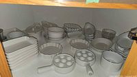 Cabinet shelf showing majority of white ceramic dishes including stacked rectangular bakers, fluted tart pans, handled bowls, escargot plate