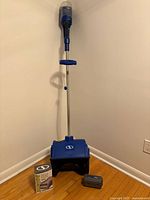Front view of assembled blue Snow Joe cordless snow shovel standing upright with battery and charger box on floor