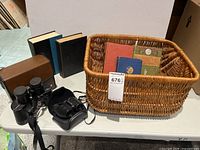 Table with Bushnell binoculars, Nikon binoculars, four hardback books upright, wicker basket holding additional books