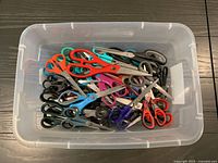 Top view of clear plastic bin filled with assorted scissors of various colours and sizes