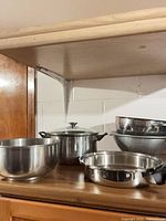 Stainless mixing bowl, stockpot with lid, colander nested with bowl, shallow frying pan on wooden shelf
