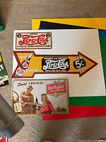Three metal advertising signs: two Pepsi-Cola (rectangular and arrow) and one Dr Pepper (rectangular) laid on coloured boards