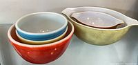 Stacked red, yellow, blue, white Pyrex mixing bowls beside tan oval casserole with white insert