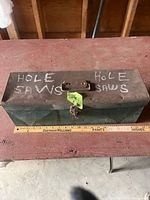 Closed green metal toolbox labelled HOLE SAWS beside yardstick showing 19 in length