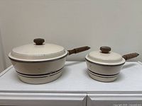 Two cream enamel West Bend saucepans with lids showing size difference and exterior stripe detail