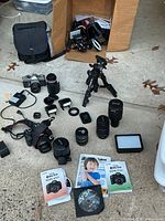 Full contents of camera lot laid out showing DSLR, film SLR, multiple lenses, flashes, tripod, LED light, manuals and accessories