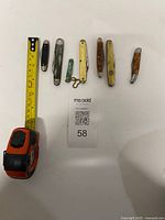 Seven pocket knives laid out with tape measure for scale