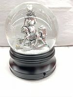 Front view of glass snow globe showing nutcracker on rocking horse and gifts inside, black base visible