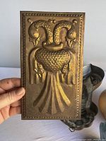 Rectangular embossed metal plaque with double-headed swan motif held in hand