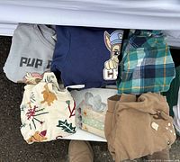 Assorted 2T shirts and pants laid out showing graphics, plaid and holiday prints