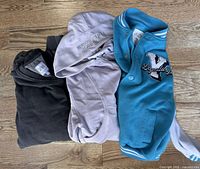 Three youth sweaters laid flat on floor: dark grey hoodie, light lavender zip hoodie, teal varsity jacket