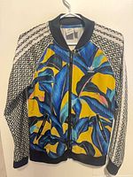 Front view of Adidas Farm Rio track jacket showing tropical print, zipper and pockets