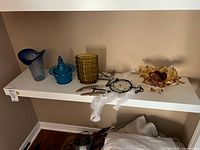 Shelf view showing blue vase, blue candy dish, stacked amber glasses, dreamcatcher, amber art glass bowl