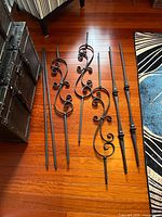 All eight matte black metal stair balusters laid on wood floor showing decorative and straight designs