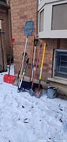 Group of shovels, skimmer, broom, watering can outside against brick wall