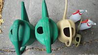 Top view of two large green watering cans, one medium tan watering can, one small tan watering can, and two white spray bottles