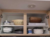 Cabinet shelf showing cream mixing bowl, casserole in metal rack, white floral serving bowl, wood bowl, wicker basket