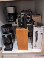 Cabinet view showing Krups coffee maker, Sunbeam toaster, knife block with knives, sharpening steel, Shardor grinder, KitchenAid hand blender box