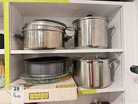 Shelf showing two stainless steel pots on top shelf, springform pans, glass platter, boxed tray, large stock pot on lower shelf