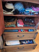 Cabinet shelves and drawers with scarves, hats, visor, crossbody bag and folded clothing visible