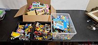 Overview of cardboard box, two black trays of LEGO pieces and clear bin with manuals