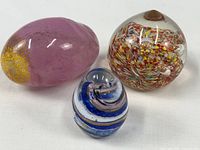 Group shot of three glass pieces: pink egg, confetti swirl paperweight, blue swirl egg