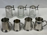 Group shot showing all seven vessels: three lidded glass steins rear, four pewter tankards front