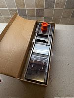Mandoline slicer in cardboard box showing stainless steel body and orange safety guard