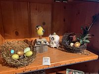 Shelf view showing two twig nests, wooden base with bird figure and ceramic birdhouse