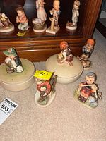 Overall lot view showing several Hummel figurines and two round lidded boxes on carpet