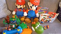 Entire lot displayed on couch: two Mario plush, LEGO box, Yoshi head, playset pieces and figures