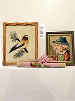 All three textile artworks on shelf: framed bird hooking, framed man petit point, rolled cross-stitch
