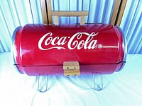 Front view showing full red barrel grill with Coca-Cola logo, wood handle, latch, and legs